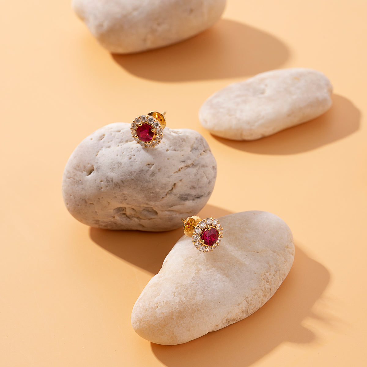 Ruby and diamond halo stud earrings in 18K gold beside a river stone