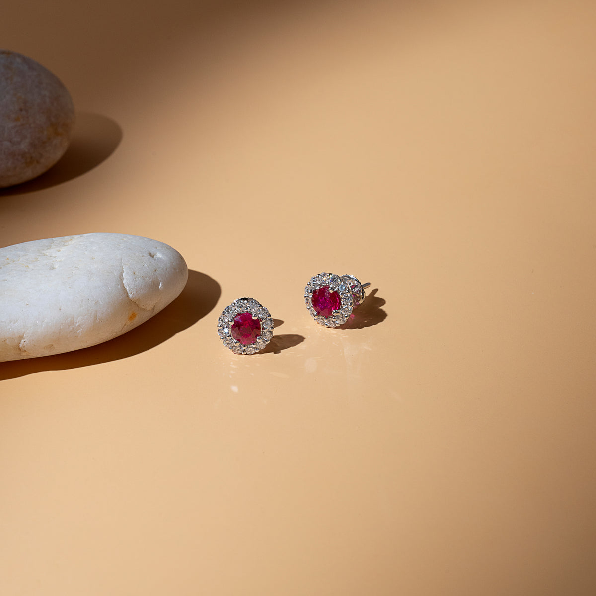 Ruby and diamond halo stud earrings in 18K gold beside a river stone, close view