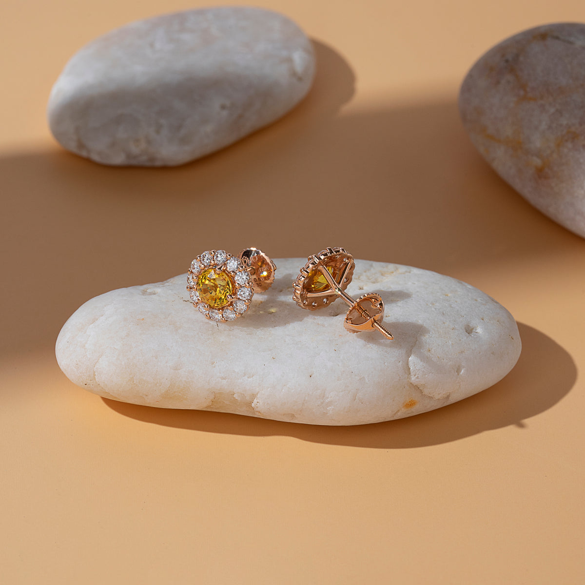 Yellow and orange sapphire heart halo stud earrings in 18K gold on a white pebble