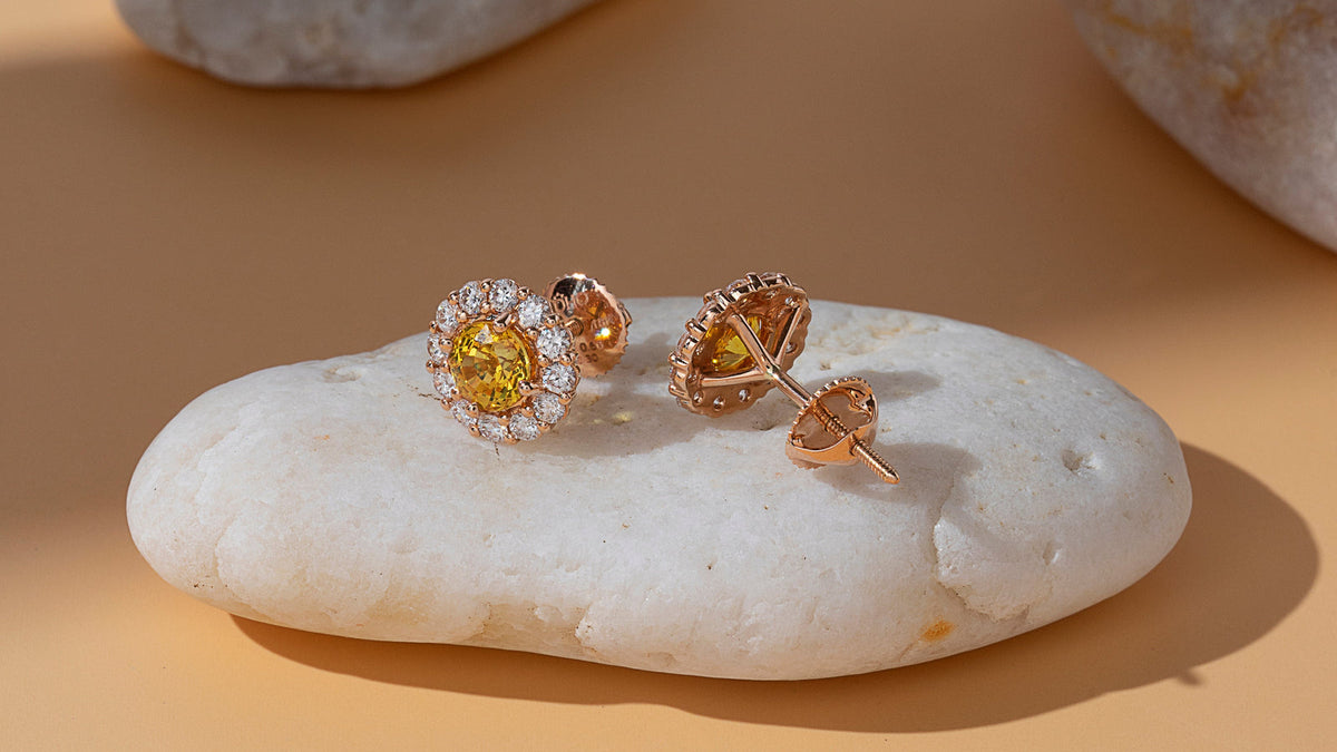 Yellow and orange sapphire heart halo stud earrings in 18K gold on a white pebble