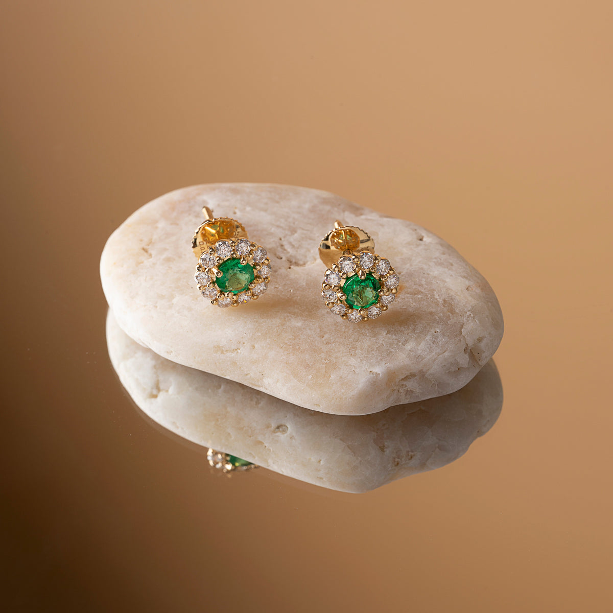 Emerald and diamond halo stud earrings in 18K yellow gold on a river stone