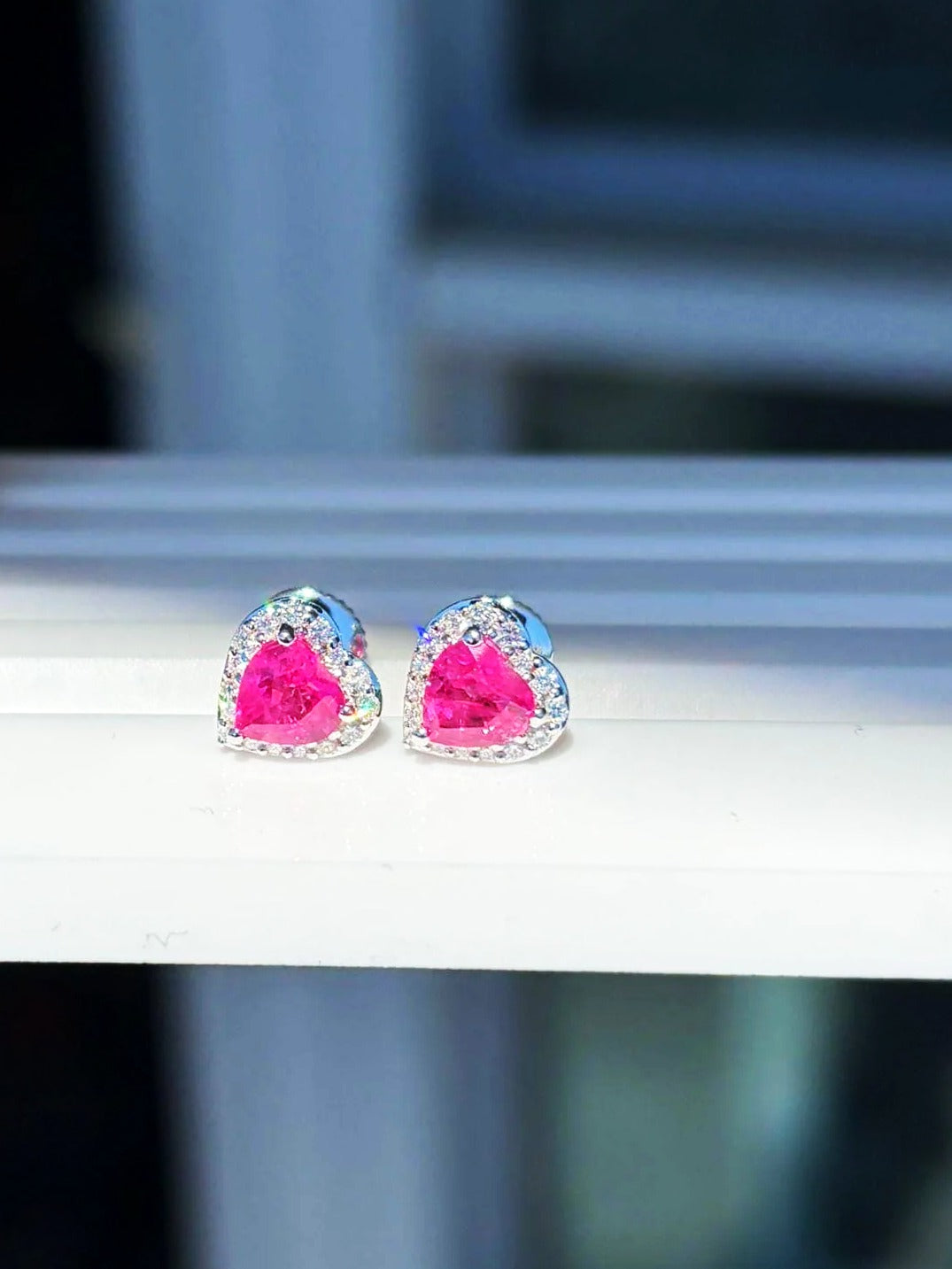 Pink sapphire and diamond heart halo stud earrings in 18K white gold on a white display stand
