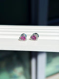 Small pink sapphire and diamond heart studs in 18K white gold on a white display stand by a window